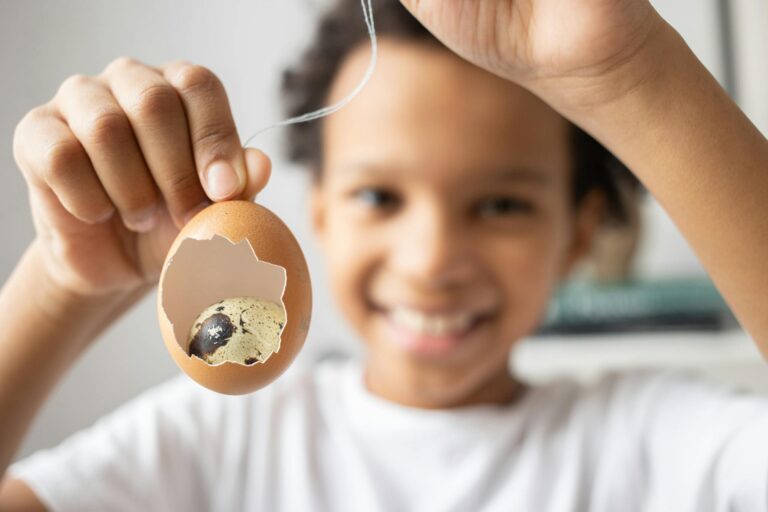 A child smiling while holding a broken egg revealing a smaller quail egg inside.